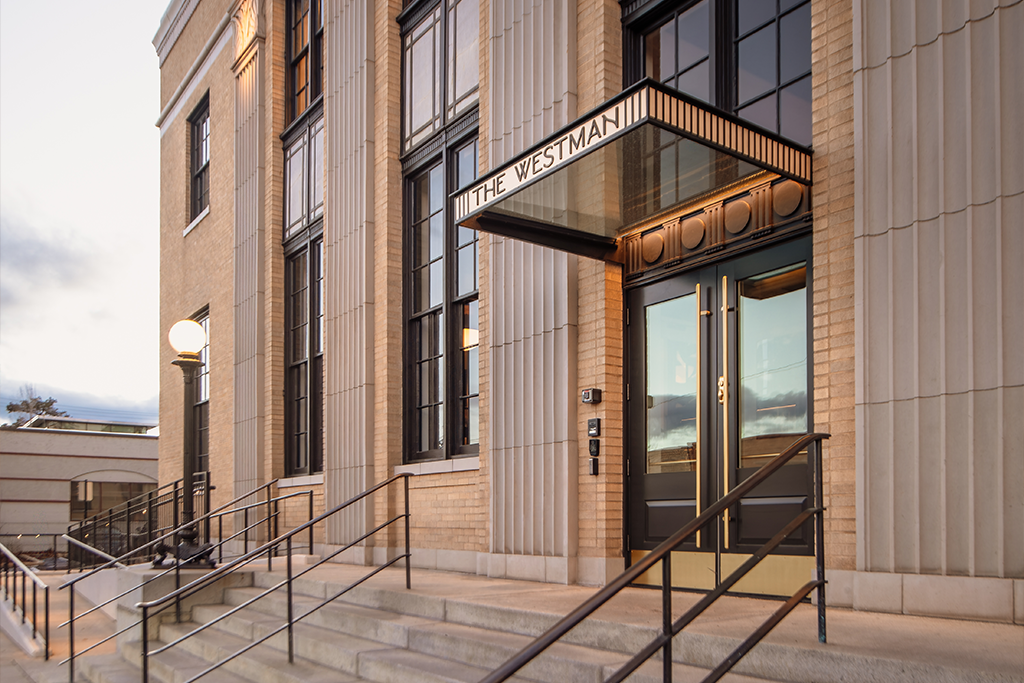Entrance of a building with tall columns, glass doors, and an awning labeled "The Westman." Stair railings and round light fixtures line the steps leading to the entrance.