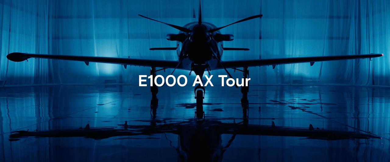 A single-engine airplane is shown from the front in a blue-lit hangar with the text "E1000 AX Tour" overlaid.
